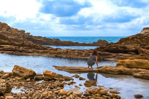 Woman crouching by a rocky tide pool on a rugged coastline, exploring the shallow water with the