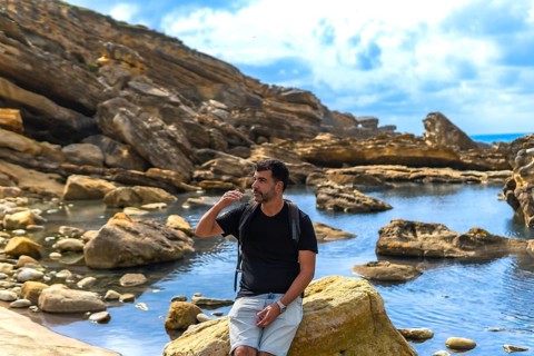 Man with a backpack sitting on a coastal rock, looking to the side and shielding his eyes from the