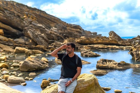 Man in outdoor clothing and backpack exploring a rugged coastal area with geological formations and