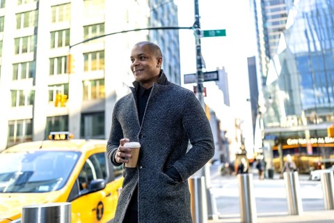 Confident black professional in smart casual coat holding a takeaway coffee cup, walking a busy