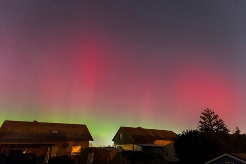 Northern lights in the night sky above houses, shimmering red and green, accompanied by sparkling