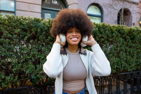 Smiling black woman with large afro hair wearing earmuffs and a sweater, standing outdoors on a
