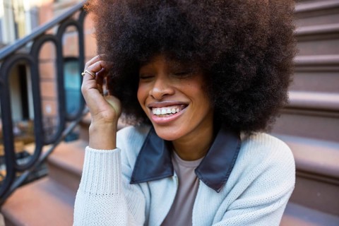 Young black woman laughing with eyes closed and hand in hair, sitting on a brownstone stoop,
