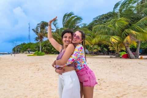 Two happy young women friends embracing on a tropical sandy beach with palm trees and a clear blue