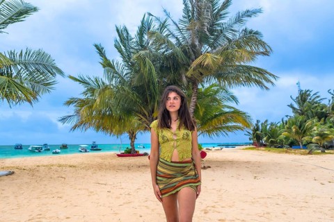 Woman standing on a sandy beach looking out towards the turquoise ocean, surrounded by lush palm