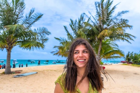 Young woman smiling and looking away, standing on a sandy beach with palm trees and a turquoise