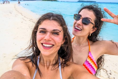Two smiling young women taking a self portrait on a white sandy beach with turquoise ocean in the
