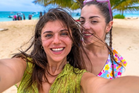 Two young happy women smiling for a close up selfie, standing on a sandy beach during a summer