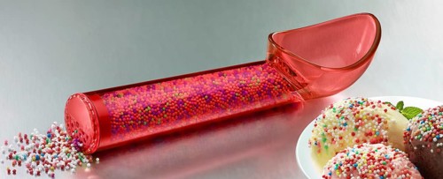 Red, transparent ice cream scoop filled with coloured sugar sprinkles, next to a plate with