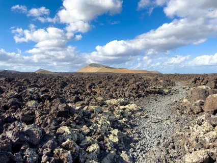 Hiking trail through lava rock to Caldera Blanca volcano against blue sky with white clouds, Mancha