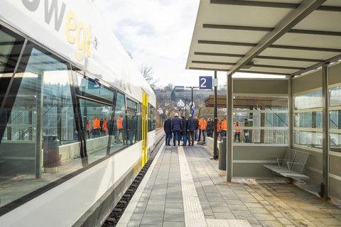 People standing in a group on a platform next to a train at a bus stop, the Hermann Hessebahn ride,