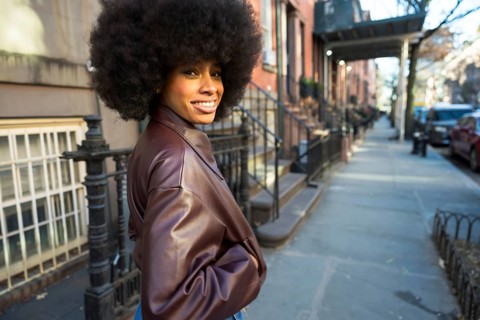 Black woman with an afro hairstyle smiling while walking on a sidewalk in front of brownstone