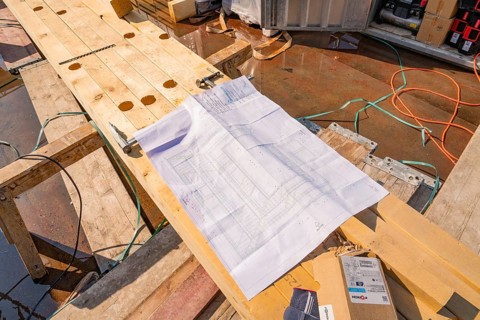 A construction plan is lying on wooden planks on a construction site, surrounded by cables and