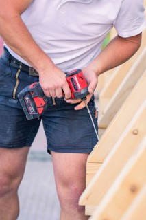 A construction worker in shorts holds a screwdriver and works on a wooden structure, carpentry, new