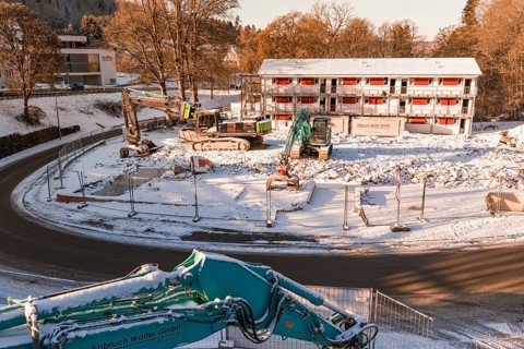 Snow-covered construction site with several excavators and a large building in the background,