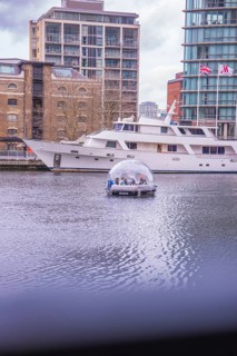 A dome boat on calm water in front of a modern marina with buildings in the background, London,