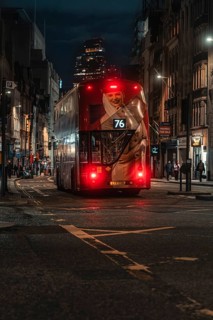 Double-decker bus travels at night through an illuminated city street surrounded by buildings,