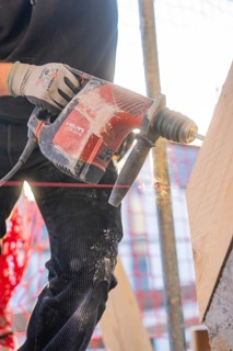A worker is drilling wood with a red electric drill on a construction site. Dust flies up,