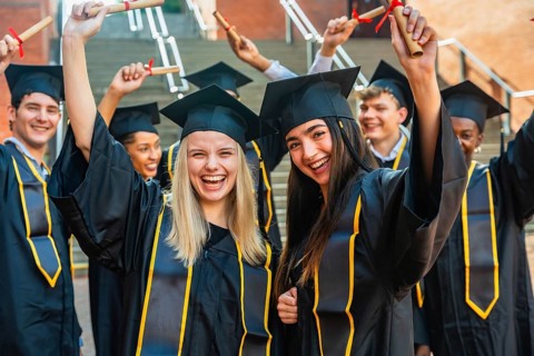 Group of happy student graduates wearing caps and gowns, cheering and holding rolled up diplomas,