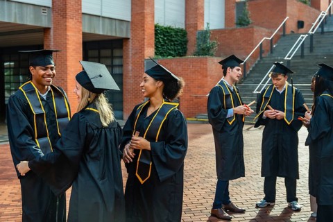 Happy diverse students wearing graduation caps and gowns, holding diplomas, and conversing outdoors