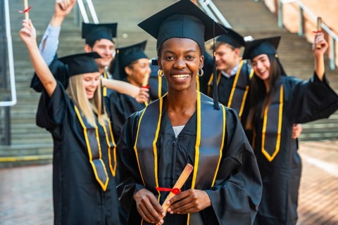 Diverse group of smiling young graduates in caps and gowns on campus, holding diplomas and
