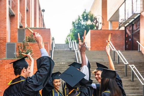 Group of diverse graduating students wearing academic caps and gowns, happily raising their diploma