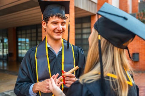 Graduating male student holding a rolled diploma, smiling and talking with a female classmate, both