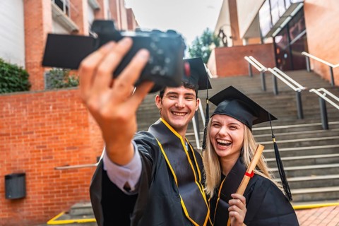 Graduating young man and woman wearing academic dress holding a diploma and filming a celebratory