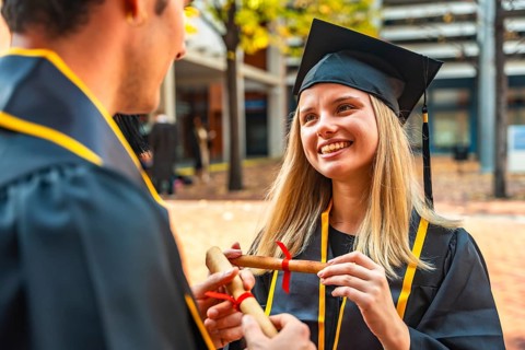 Smiling university graduate in cap and gown accepts rolled diploma from unseen hand, celebrating