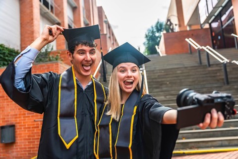 Two cheerful university graduates, a man and a woman, wearing cap and gown, smiling and making