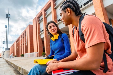 Two diverse young students engaging in a friendly conversation while sitting on a ledge on the