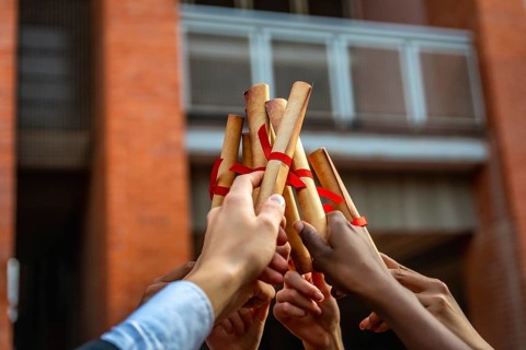 Graduates from various backgrounds raise their rolled up diplomas, tied with red ribbons,