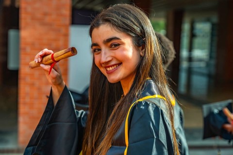 Smiling young woman celebrating graduation, proudly holding her rolled up diploma, wearing a cap