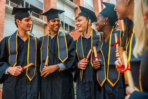 Joyful group of diverse university students wearing graduation caps and gowns, holding rolled