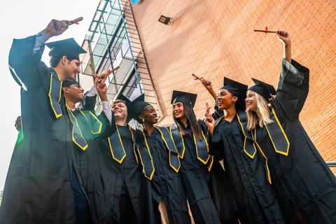 Diverse group of joyful young graduates in black caps and gowns celebrating on campus, holding