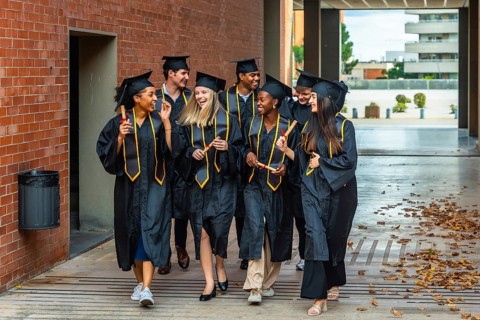 Diverse group of happy graduates celebrating achievement, walking on campus wearing academic gowns,
