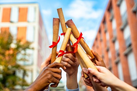Diverse hands are holding rolled diplomas tied with red ribbons, raising them up in celebration of