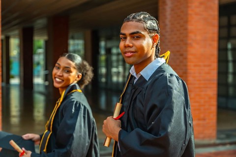 Two diverse university graduates smile and pose on campus in caps and gowns, proudly holding