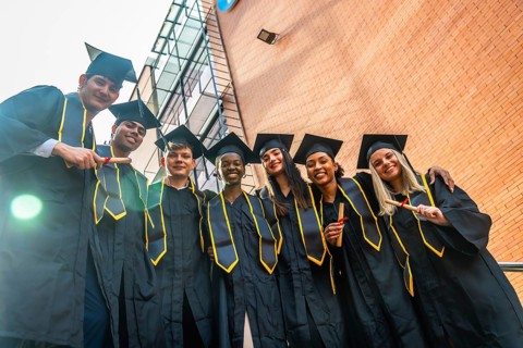 Diverse group of joyful university graduates in caps and gowns standing outdoors by campus