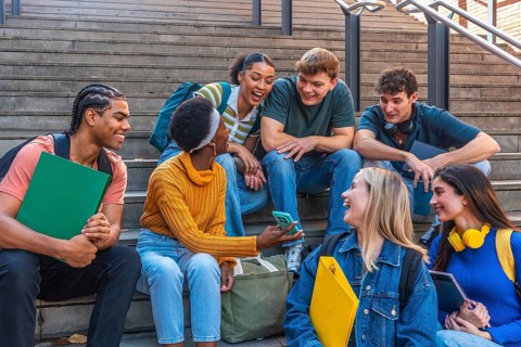 Group of young multi ethnic students sitting on steps, laughing and looking at a smartphone,