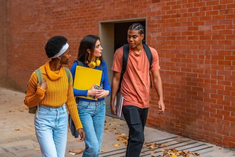 Three diverse young university students walking along a brick wall, carrying backpacks and books,