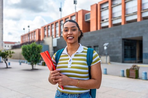 Young female student smiling while holding a folder and wearing a backpack, standing outdoors on a