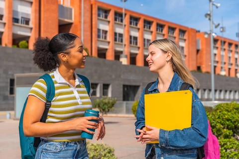 Two diverse female students walking on a university campus, smiling and engaged in conversation