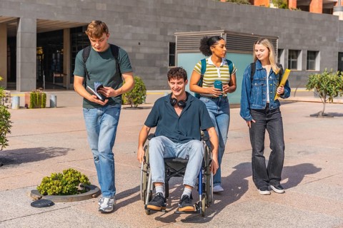Group of four diverse university students walking on campus outdoors, one young man in a wheelchair