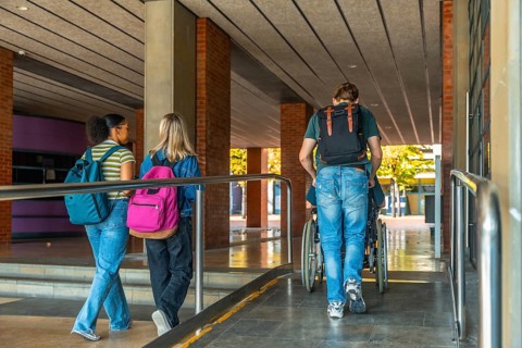 University students walking up an accessible campus ramp, one friend pushing another in a