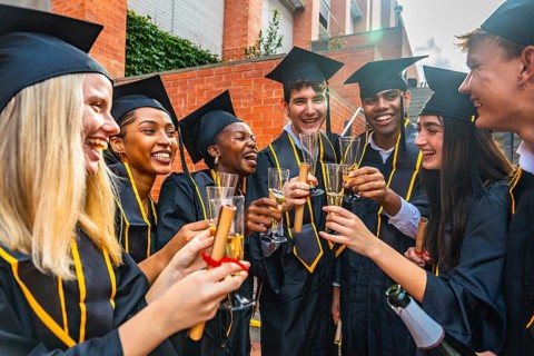 Young diverse international students wearing graduation caps and gowns toasting with champagne