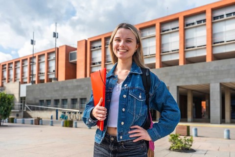 Happy young female student with backpack and folder smiling, standing in front of a modern