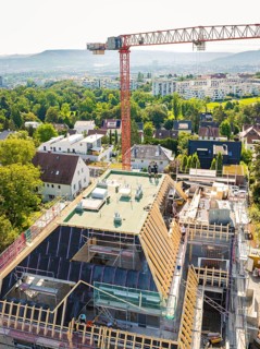 A construction site with a crane over a roof, an urban and green landscape in the background,