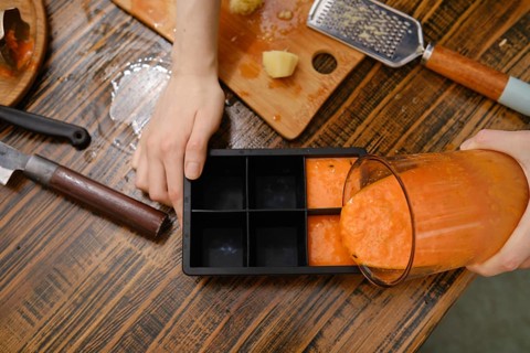 A person is pouring fruit mixture from a blender into a silicone ice cube tray. The table has