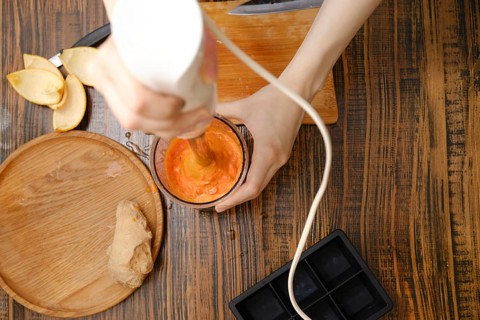A person uses a hand blender to mix fruits and ginger in a cup. The setting looks like a kitchen